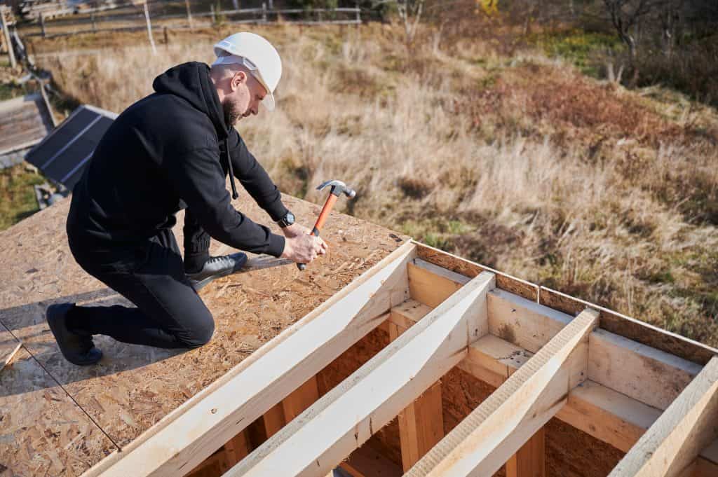 A person wearing a hard hat uses a hammer to nail down plywood on a roof under construction, with exposed wooden beams visible.