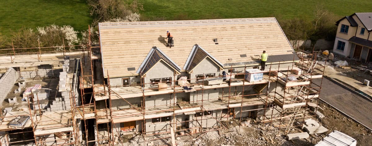 Aerial view of a partially constructed house with scaffolding, workers on the roof, and building materials scattered around the construction site.