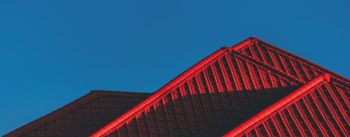 Red tiled rooftops with triangular peaks are shown against a clear blue sky, with sunlight casting shadows across the tiles.