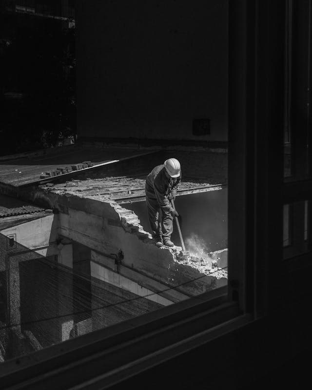 A construction worker wearing a hard hat is demolishing a concrete rooftop with a tool, viewed through a window in black and white.