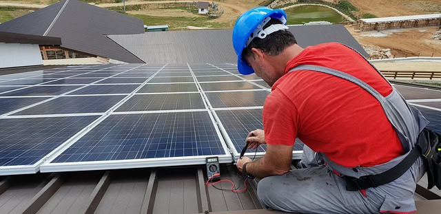 A worker in a blue helmet and red shirt uses a multimeter to check solar panels installed on a rooftop.