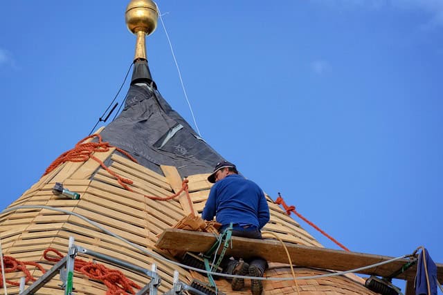 A construction worker is sitting on scaffolding, repairing the wooden shingles on a conical rooftop with ropes and tools, under a clear blue sky.
